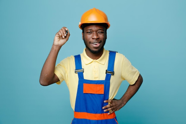 Showing tongue putting hand on hips young african american builder in uniform holding marker isolated on blue background