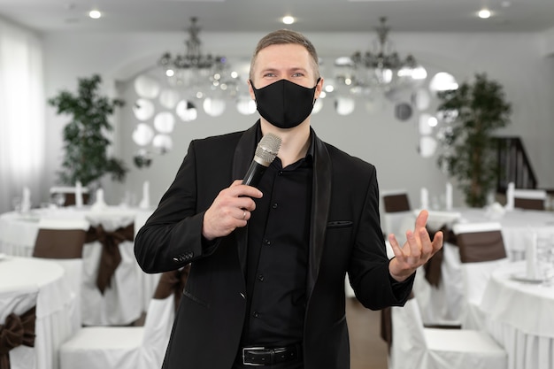 Showman in tuta e maschera protettiva con un microfono in mano nella sala banchetti di rastoran.