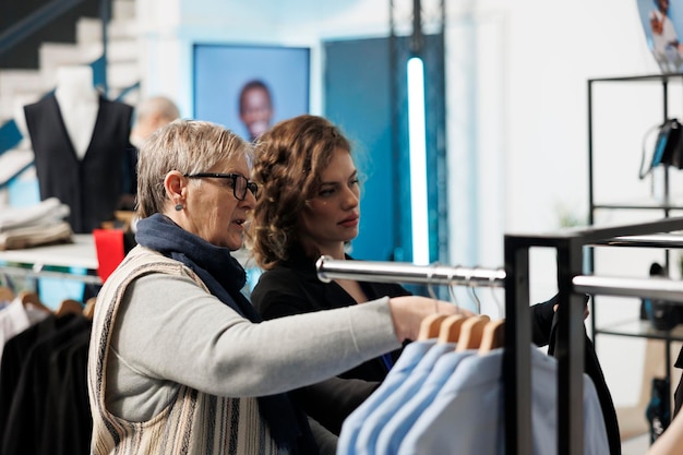 Showroom employee helping elderly woman choosing trendy clothes, discussing fabric in modern boutique. Senior client shopping for casual wear, buying fashionable merchandise in clothing store