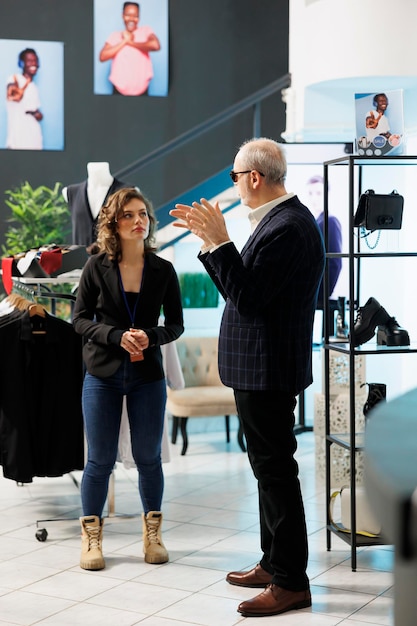 Showroom employee helping senior stylish customer choosing right sunglasses in modern boutique. Elderly client shopping for formal wear, buying fashionable clothes and accessories in clothing store