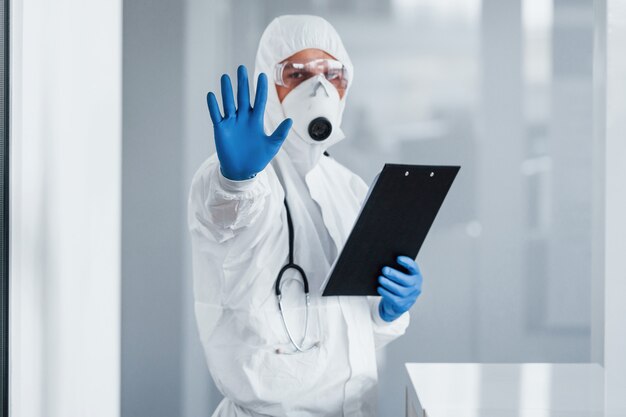 Shows stop gesture by hand. Male doctor scientist in lab coat, defensive eyewear and mask holding notepad in hands