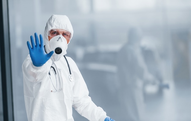 Shows stop gesture by hand. Male doctor scientist in lab coat, defensive eyewear and mask