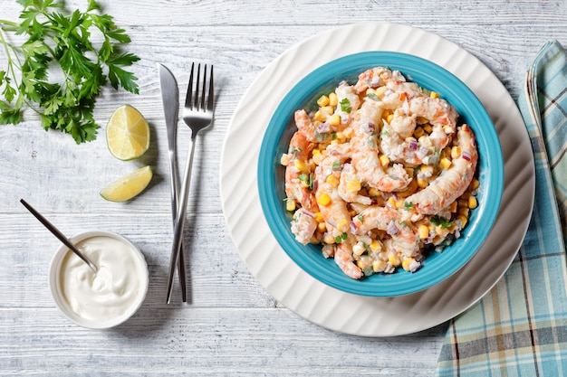 Shrimp salad with sour cream and lime juice dressing, corn, red onion, and parsley served on a blue salad bowl on a white charger plate with cutlery on a white wooden background, top view