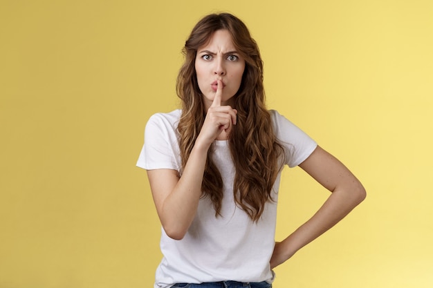 Shut your mouth no talking during concert. Serious-looking strict displeased adult sister demand sit silent hushing hold index finger pressed lips frowning angry shushing yellow background