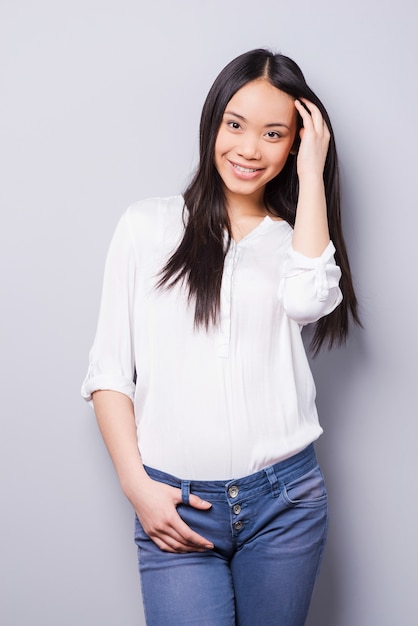 Shy beauty. Beautiful young Asian woman looking at camera and holding hand in hair while standing against grey background