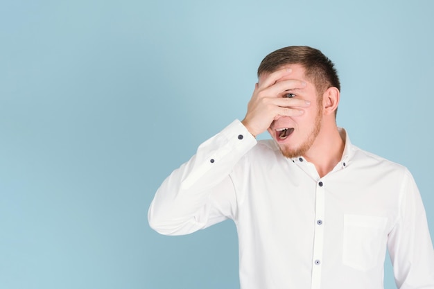 A shy young adult man with a beard peeps through his fingers covering his face with his palm Copy space