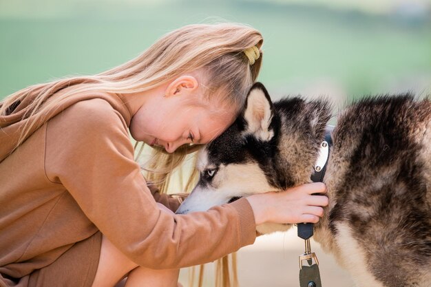 Siberian husky and his little girl owner hug together on the road against the backdrop of a rural landscape in summer