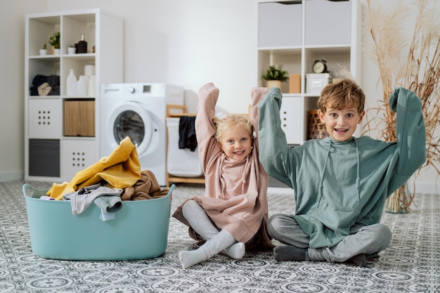 The siblings are sitting on floor in clean laundered\
sweatshirts taken from bowl of clothes