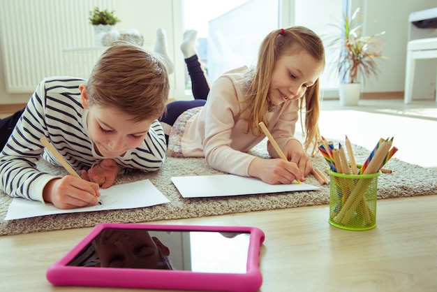 Siblings drawing over paper by digital tablet while lying at home