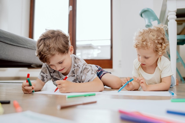 Siblings drawing with colored markers while lying on wooden floror at home in the living room