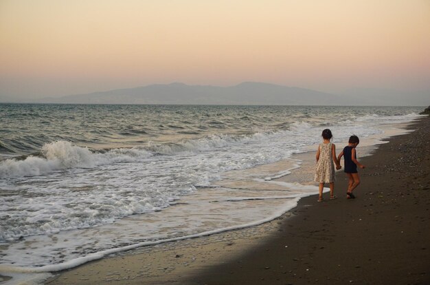 Photo siblings holding hands while walking at beach against sky during sunset