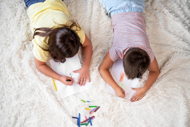 Foto fratelli che giocano insieme a casa vista dall'alto di un ragazzino e una ragazza sdraiati sul tappeto e disegnando su fogli di carta bianchi con pastelli colorati