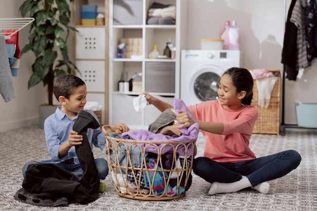 Siblings sit on the laundry room floor sorting colored clothes before putting them in the machine