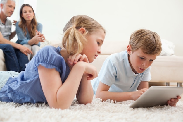 Братья и сестры, использующие планшетный компьютер с родителями на заднем плане