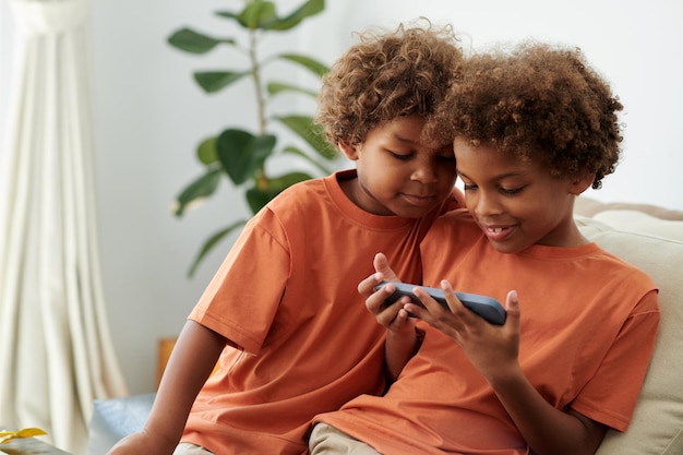 Siblings watching entertaining video on smartphone when sitting on couch at home