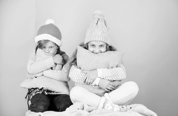 형제 자매는 겨울 따뜻한 모자를 입고 분홍색 배경에 앉아 있습니다. 어린이 소년과 소녀는 베개와 모자로 몸을 따뜻하게합니다. 따뜻하고 편안하게 지내십시오. 귀엽고 아늑한 액세서리로 겨울 옷을 따뜻하게하십시오