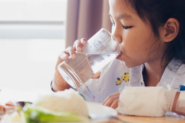 病院の個室に滞在中に一人で昼食のために健康的な食べ物を食べた後、コップ一杯の水を飲む病気のアジアの子供の女の子。ヘルスケアとライフスタイルの概念。