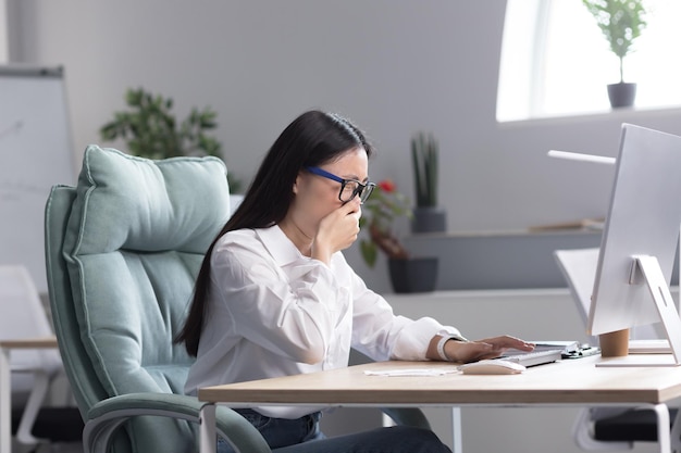 Sick asian female office worker has cold and flu coughs and sneezes into tissue works at computer