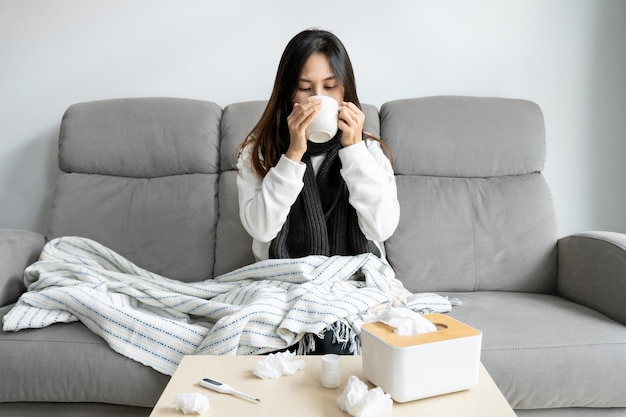 インフルエンザの症状を持つ病気のアジアの女の子は、ソファーに座って温かい水を飲み、組織薬と体温計を机の上に置きます 風邪インフルエンザと健康問題のコンセプト コピースペース