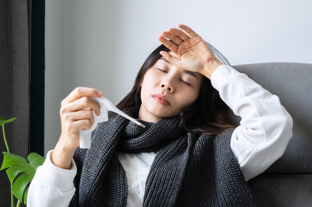 Sick Asian girl sitting on couch at home hand holding a thermometer and touching her forehead. Winter cold and flu concept. Close up