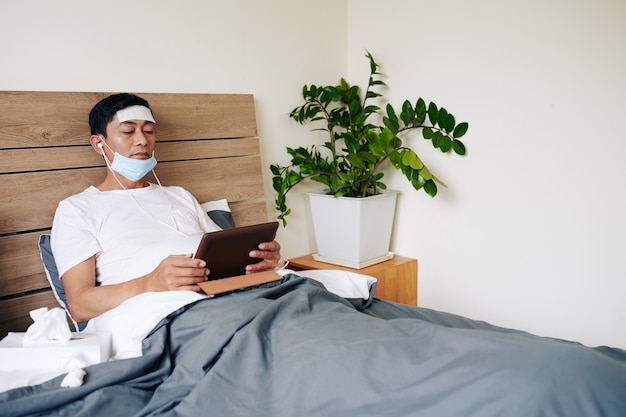 Больной азиатский мужчина с охлаждающим гелевым пластырем на лбу и медицинской маской, лежащий в постели с компьютером талбет, оставаясь дома из-за коронавирусной болезни