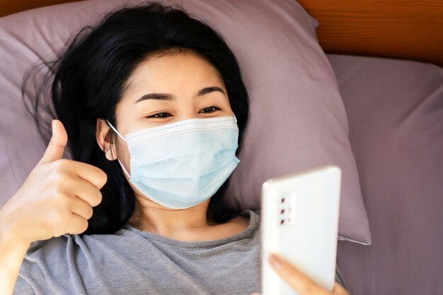 写真 スマートフォンを握っている病気のアジア人女性ベッドで手を叩く映像通話