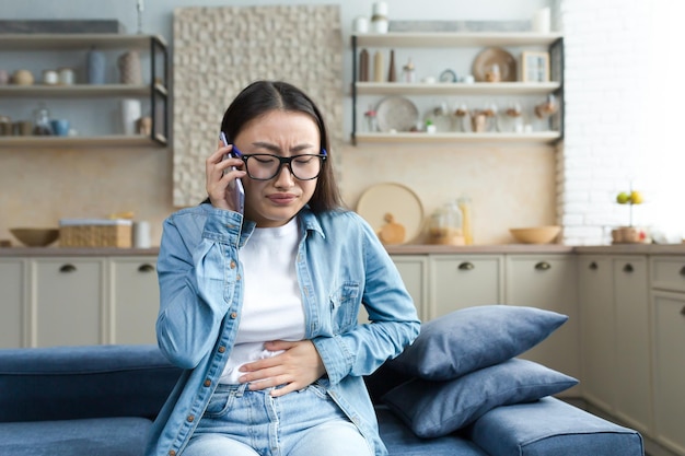 Sick asian woman at home woman sitting on sofa and having severe stomach pain calling hospital for