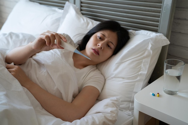 sick asian woman with flu virus lying in bed measuring temperature. upset frowning lady look thermometer. illness female resting at home hand holding tissue with medicine and water on desk beside.