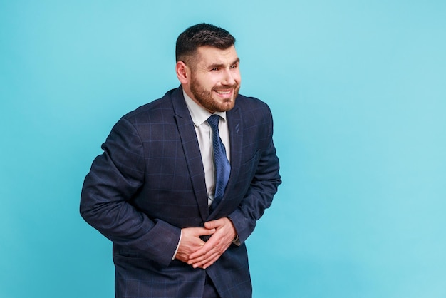 Uomo d'affari barbuto malato che indossa un abito in stile ufficiale tenendo le mani sullo stomaco sentendo dolore acuto, indigestione e nausea, ulcera duodenale. studio indoor girato isolato su sfondo blu.