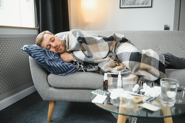 Sick bearded man who has bad cold or seasonal flu sitting on\
couch at home guy with fever wearing warm plaid shivering with\
worried face expression