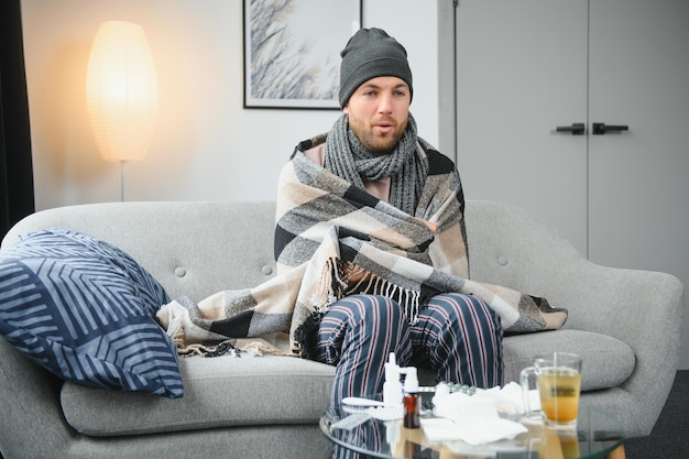 Sick bearded man who has bad cold or seasonal flu sitting on\
couch at home guy with fever wearing warm plaid shivering with\
worried face expression