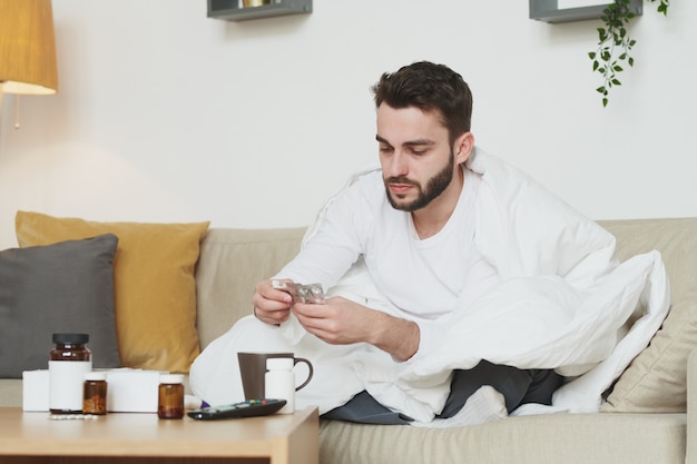 Sick bearded man with high body temperature taking pills or tablets while staying at home because of coronavirus or other illness