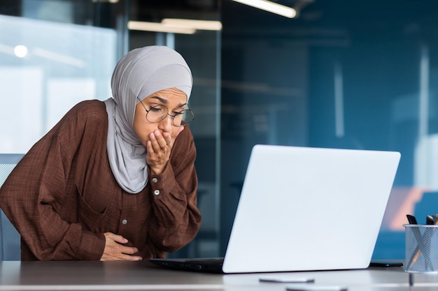 Sick business woman in hijab working inside office at workplace nausea and stomach pain in muslim