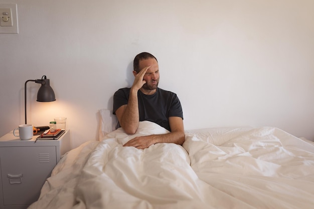 sick caucasian man spending time at home self isolating and social distancing in quarantine lockdown during coronavirus covid 19 epidemic, sitting up in bed with a pained expression, touching his head