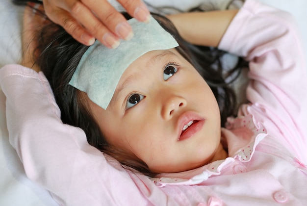 Sick child girl lying on bed with mother stick cool jell on her daughter head.