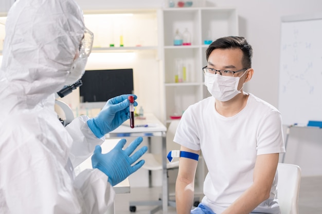 Sick chinese guy in protective mask sitting in front of lab worker in gloves and coveralls holding sample of his blood in flask