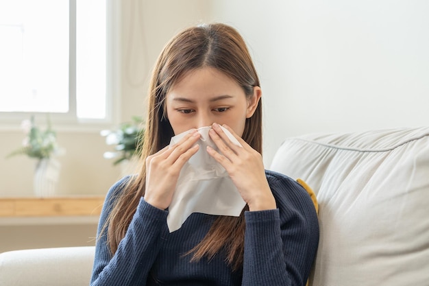 病気のコロナウイルスcovid19アジアの若い女性の女の子の毛布の下での頭痛はインフルエンザになり、自宅のソファベッドに座って鼻水をくしゃくしゃにする組織紙を使用しますウイルスの人の健康管理
