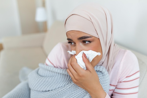 Sick day at home young arabic woman has runny and common cold\
cough closeup of beautiful young woman with hijab caught cold or\
flu illness