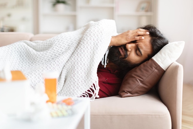 Photo sick eastern guy laying on couch covered with warm blanket