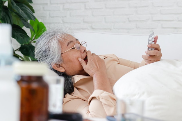 집에 있는 소파에 누워 있는 아픈 노인 아시아 여성이 병을 치료하기 위해 알약 캡슐을 들고 집에 있는 노인을 위한 건강 관리 개념