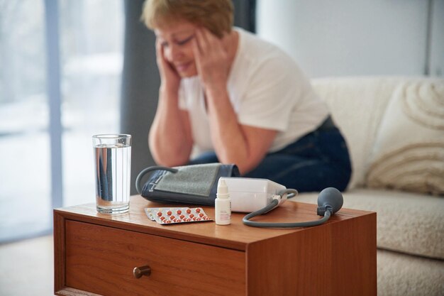 病気の気分は悪い 薬はテーブルに 高齢の女性は自宅のリビングにいます