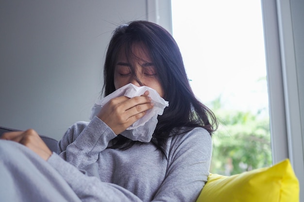 Sick girl sitting on the sofa in the house, having a runny nose fever and wiping with a sick handkerchief. cold, sneezing concept