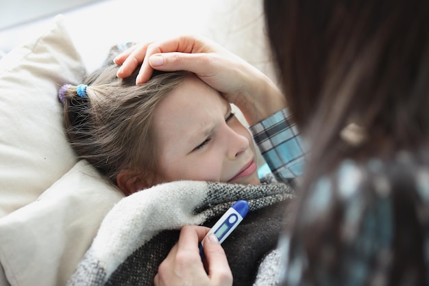 Фото Больная девочка остается в постели, мать измеряет температуру с помощью электронного термометра