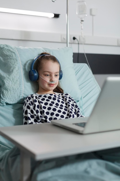 Sick girl with headphones watching funny cartoons on laptop while sitting on patient bed inside hospital pediatrics ward room. Ill kid under treatment enjoying internet video content inside clinic.