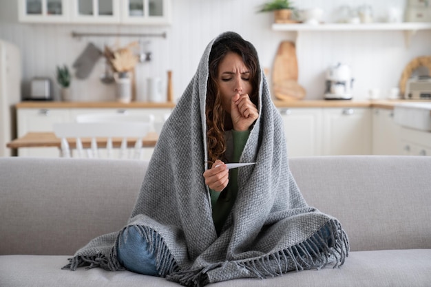 Sick at home unhealthy young woman coughing while measuring body temperature with thermometer