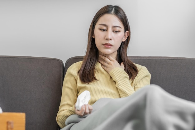 아픈 상처나 통증 인후통이 있는 아시아 젊은 여성 소녀는 발열 독감에 걸리고 티슈 페이퍼를 사용하여 집 소파 침대에 앉아 재채기 코 콧물을 흘리고 있습니다.