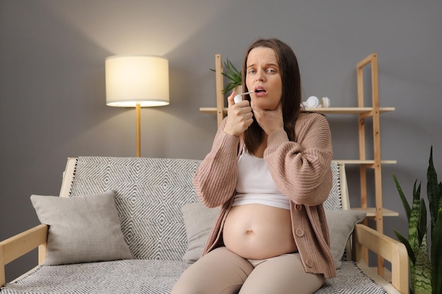 Фото Больная беременная белая женщина, сидящая дома на диване с болью в горле, использующая спрей для медицинского лечения, заботясь о своем здоровье во время беременности