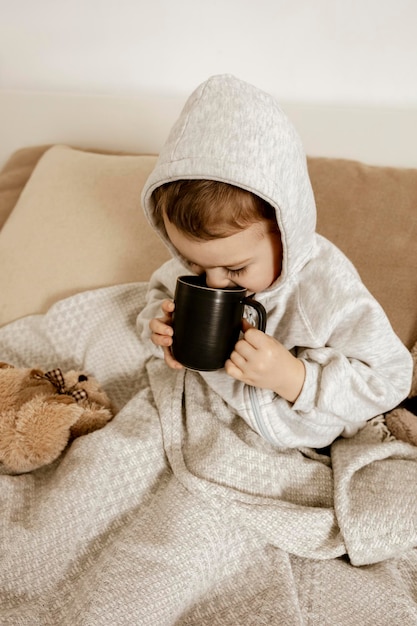 Больной маленький мальчик пьет горячий чай на кровати дома. Нездоровый, больной ребенок, завернутый в одеяло, с кружкой в своей комнате. Сезон гриппа. Интерьер и одежда в натуральных земляных тонах. Уютная обстановка.