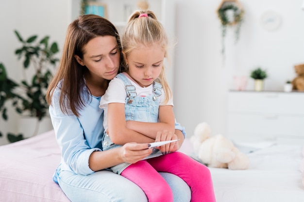 Sick little girl and her mother in casualwear looking at thermometer held by young woman while both sitting on couch