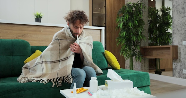 Foto uomo malato coperto da un caldo plaid che misura una temperatura e con un termometro in bocca, ha la febbre e l'influenza.
